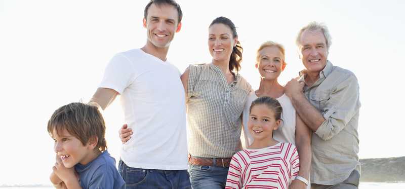 A smiling family spanning three generations standing on a beach.