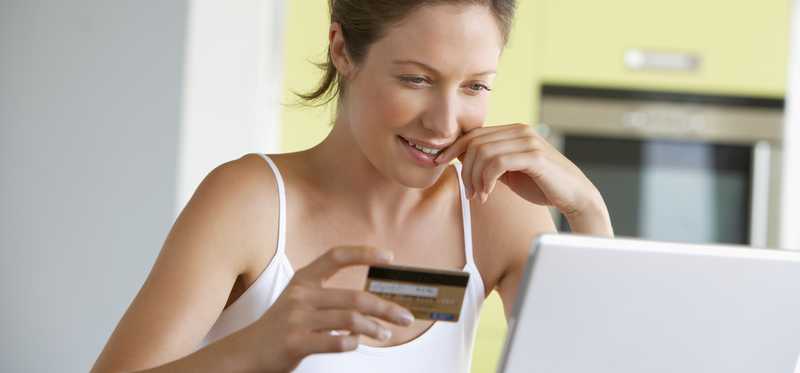 A smiling woman holding a credit card in her right hand as she makes an online purchase