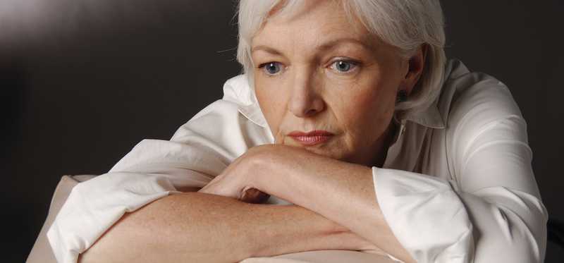 A visibly worried senior woman with her arms folded on the back of a chair and her head resting on her arms.