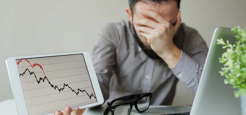 A worried investor covering his face with his left hand while holding a tablet that shows a plunging stock chart in his right hand.
