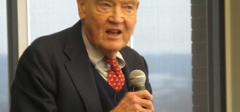 Jack Bogle sitting on a stool at Fool HQ holding a mic and speaking to a crowd.