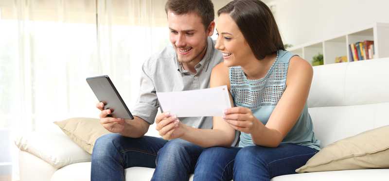 Couple sitting on couch, man holds a tablet, woman holds a check.