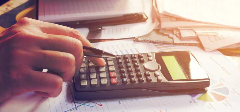 A hand using a calculator on a desk filled with paperwork.