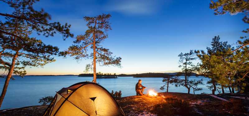 A tent pitched beside a lake at sunset and a person sitting by a campfire.