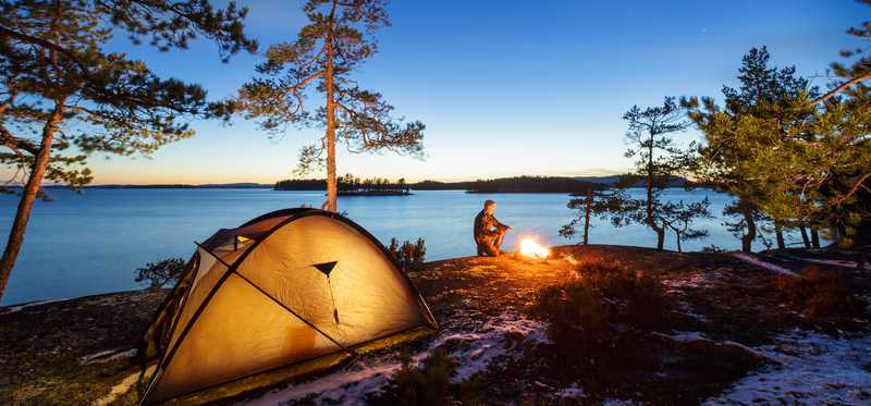 A tent pitched beside a lake at sunset and a person sitting by a campfire.