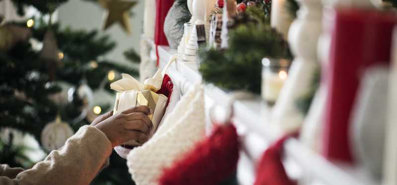 Someone's hand placing a gift into a Christmas stockings hanging on a mantel.