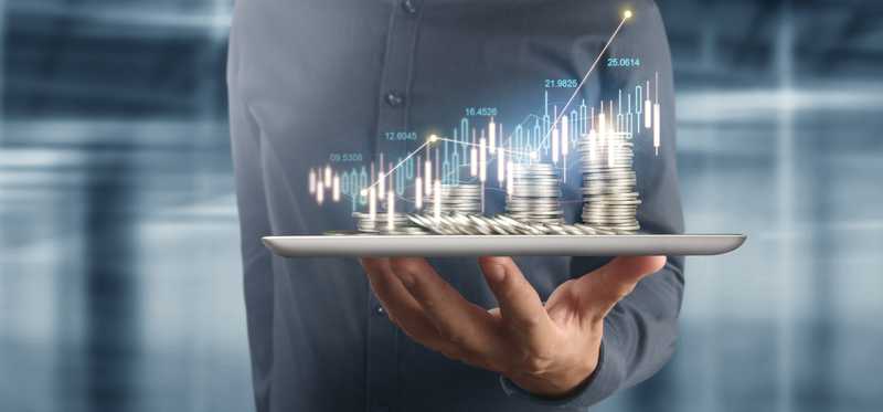 Person holding a tablet with stacks of coins sitting on top and charts in the background.