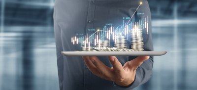 Person holding a tablet with stacks of coins sitting on top and charts in the background.