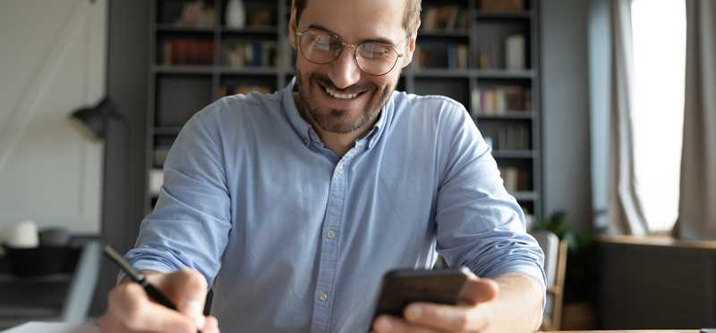 Smiling person in glasses looking at mobile phone and making notes in journal.