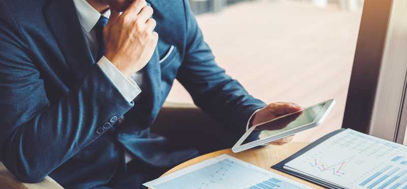Man sitting at table with tablet and printed stock charts.