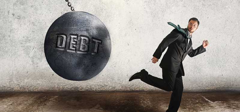 The word Debt on a wrecking ball swinging toward a person running away.