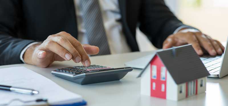 Person typing on a calculator and a laptop, with a miniature house sitting on the desk.