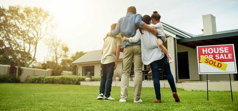 Family stands hugging in front of a home with a For Sale sign with a Sold sticker across it.