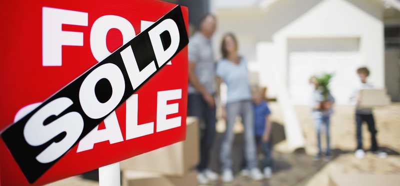 A red For Sale sign with a black Sold sticker over it in front of a house with a family moving boxes.