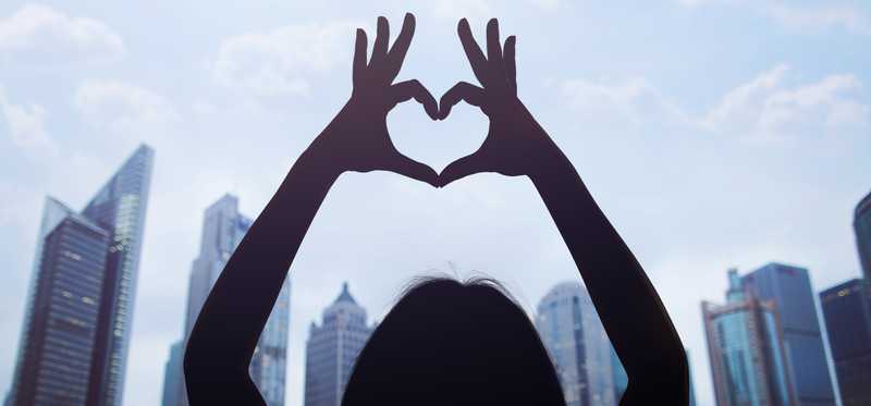 Person making heart with hands in front of city skyline.
