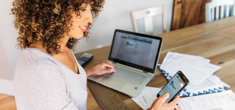 Person working on laptop and cell phone with papers at desk.