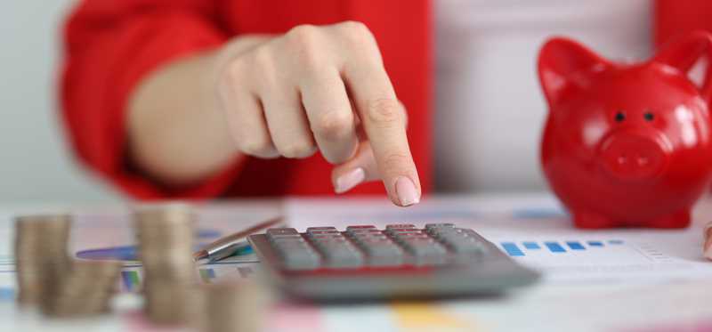 Person using a calculator at a desk with a red piggy bank, charts, and stacks of coins sitting on it.