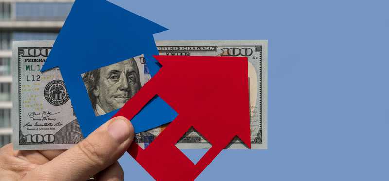 Hand holding money with blue and red paper cutouts of houses on top.
