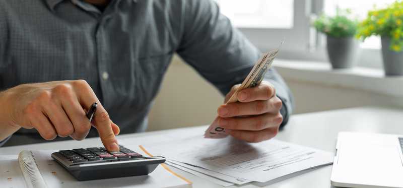 Person using calculator while holding cash and looking at papers on a desk.