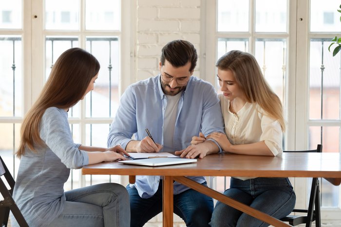 Happy young clients signing paper contract with professional broker or realtor, after discussing agreement details. Confident saleswoman watching millennial couple putting signature, purchasing house.