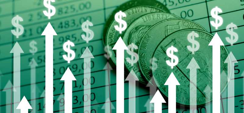 Arrows in a bar graph, pointing upward, topped with dollar signs and with coins and charts in the background.