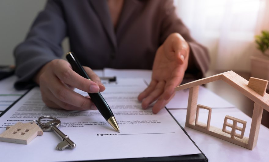 Person is sitting across a desk and using a pen to show you where to sign on a document that is sitting next to a house key and miniature home.