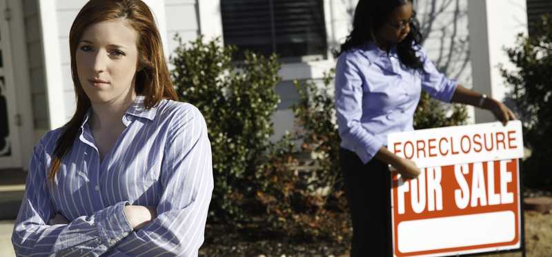Two people stand in front of a house with a foreclosure sign.