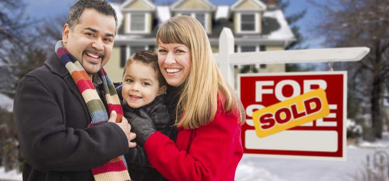 Smiling family standing next to a For Sale/Sold sign in a snowy front yard.