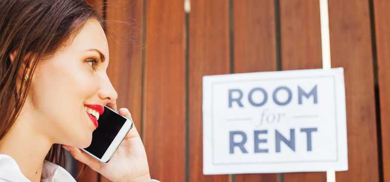 Person talking on phone in front of a fence with a sign hanging on it that says Room for Rent.