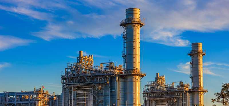 Natural gas-fired turbine power plant with its cooling towers rising into a cloud-filled blue sky.
