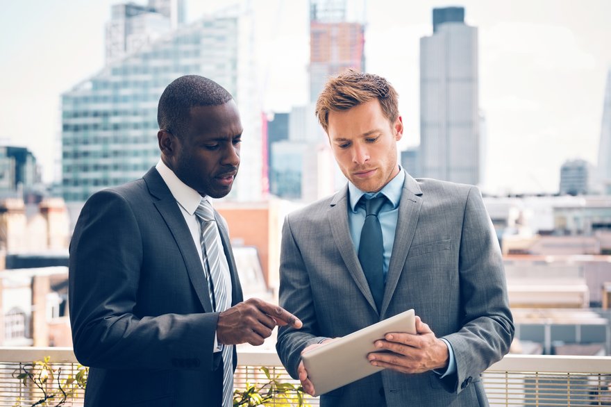 Businesspeople discussing a project on a digital tablet while standing on a balcony with a cityscape in the background.