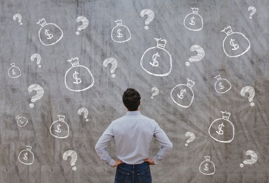 A man stands in front of a blackboard with several question marks and dollar symbols.