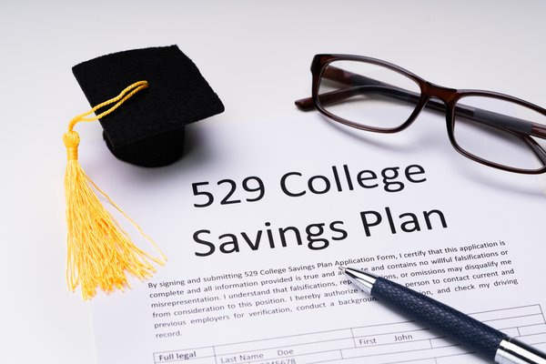 A tiny graduation cap, glasses, and pen next to a paper that reads 529 College Savings Plan.