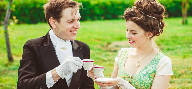 Two people dressed as aristocrats might have dressed 100 or more years ago and having tea and smiling.