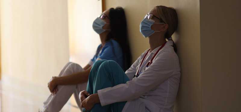 Exhausted healthcare workers are slumped against a wall.