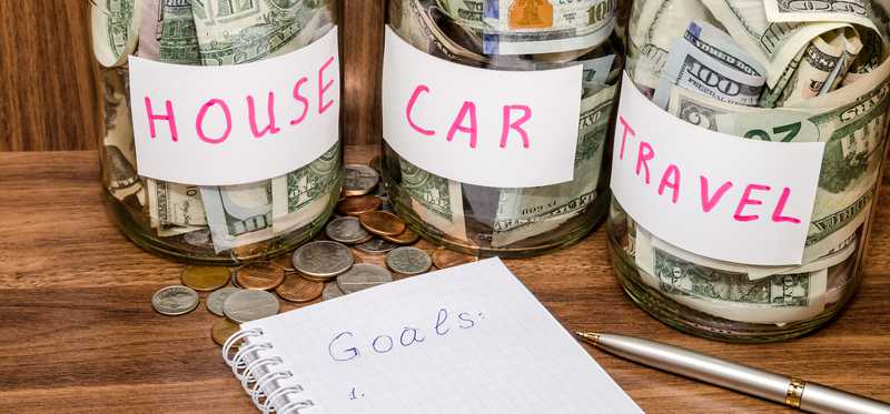 We see three savings jars full of cash and labeled house, car, and travel. Next to them is a notebook with the word goals printed in it.