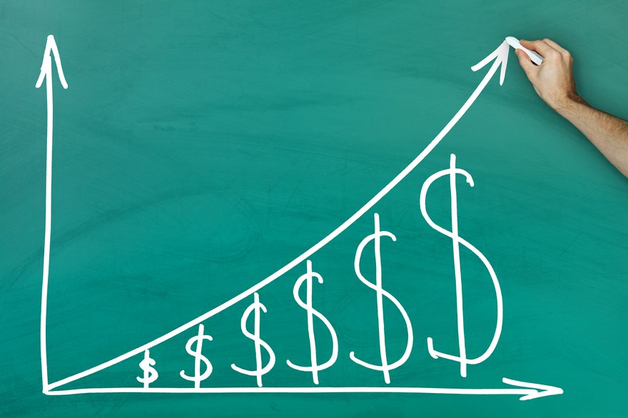 A hand is drawing a graph on a blackboard of an upward trending arrow over a series of progressively bigger dollar signs.
