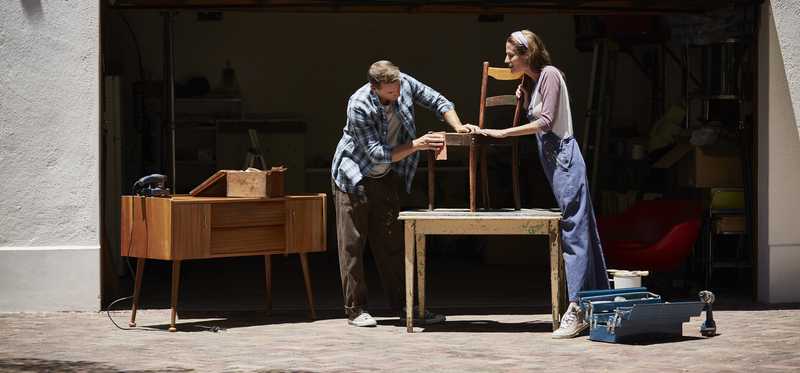 Two people are refinishing furniture by a garage.