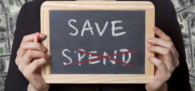 We see hands holding a small chalkboard on which is written the words spend and save, with spend crossed out