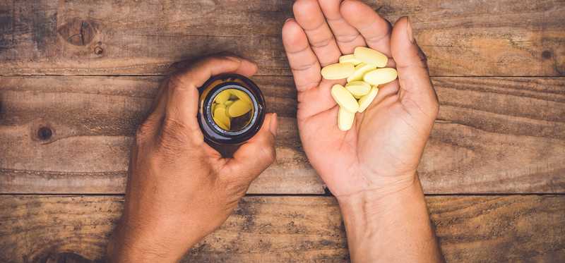 Hands lying on a wooden table and holding some pills in one hand and an open pill bottle in the other.