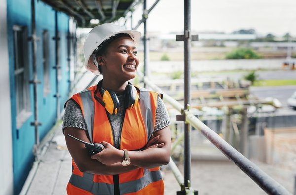 Smiling person wearing a hard hat and safety vest at a construction site.