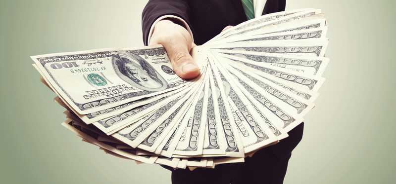 man in business suit holding out handful of hundred dollar bills
