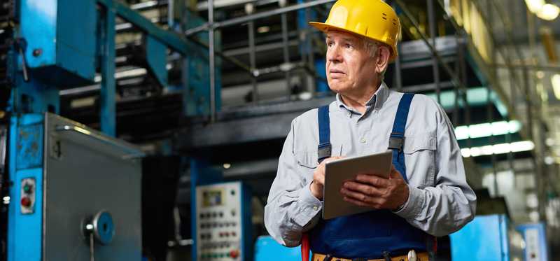 Person wearing construction hat and holding clipboard in factory.