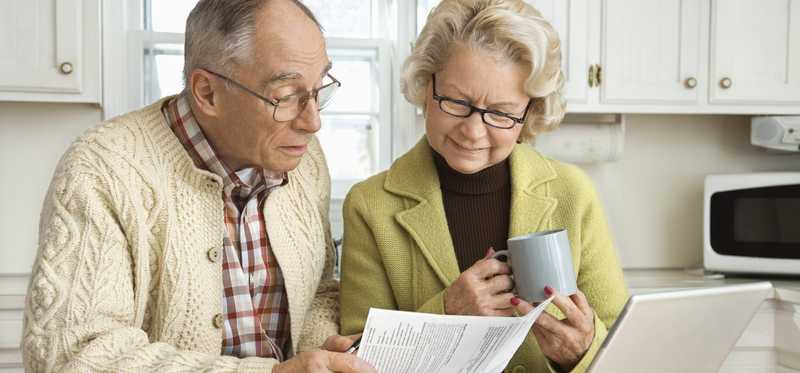Two people sitting at kitchen table and looking at papers in front of laptop.