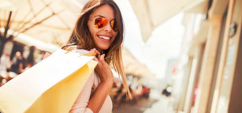 A young woman in sunglasses holds shopping bags over her shoulder.