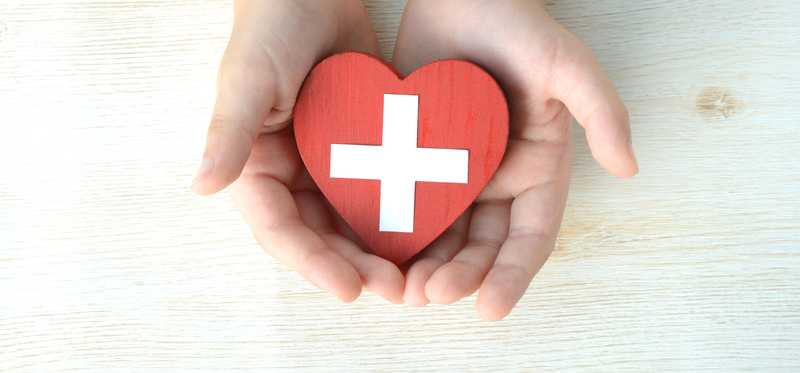 Hands holding a red heart with white cross on it.