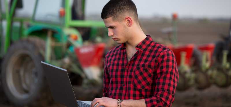 A person stands by a tractor on a farm and looks at a laptop.