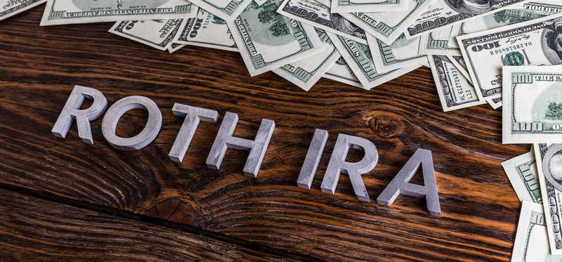 Silver letters spelling Roth IRA on floor with scattered hundred dollar bills.