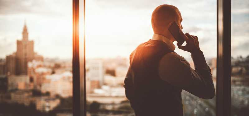 Businessperson looking out an office window at the city while talking on the phone.