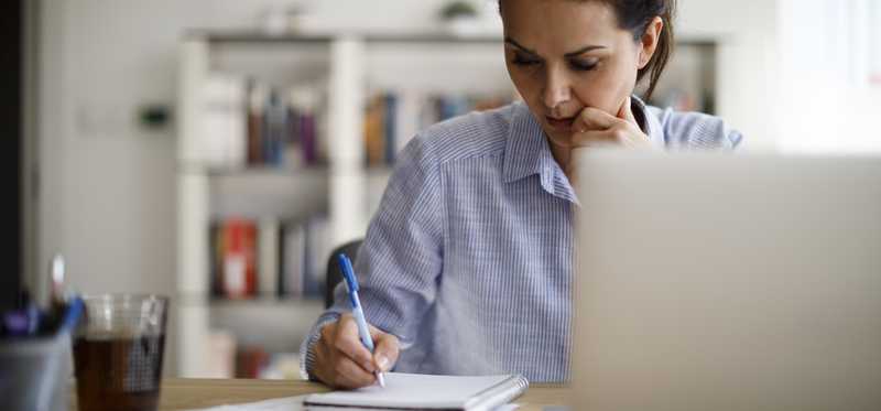 Person sitting at table with laptop open and writing in notebook.